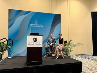 Jason Bole and Cindy Berner sit on stage smiling at the camera before presenting a breakfast session at ACLM 2024
