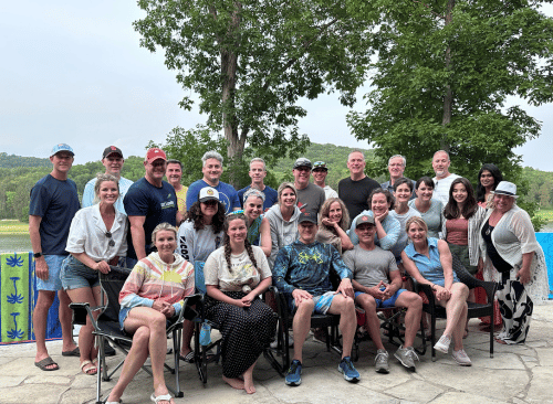 Just under 30 employees pose for a picture lined up in three tired rows with the first row sitting. Behind the group is a lake and trees.