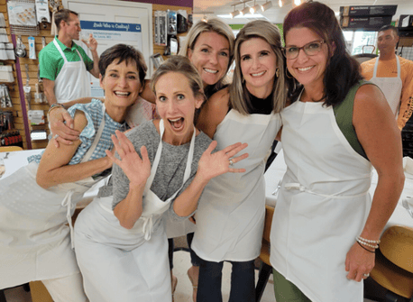 five women in white aprons smiling at the camera. All are huddled together with their arms around each other 