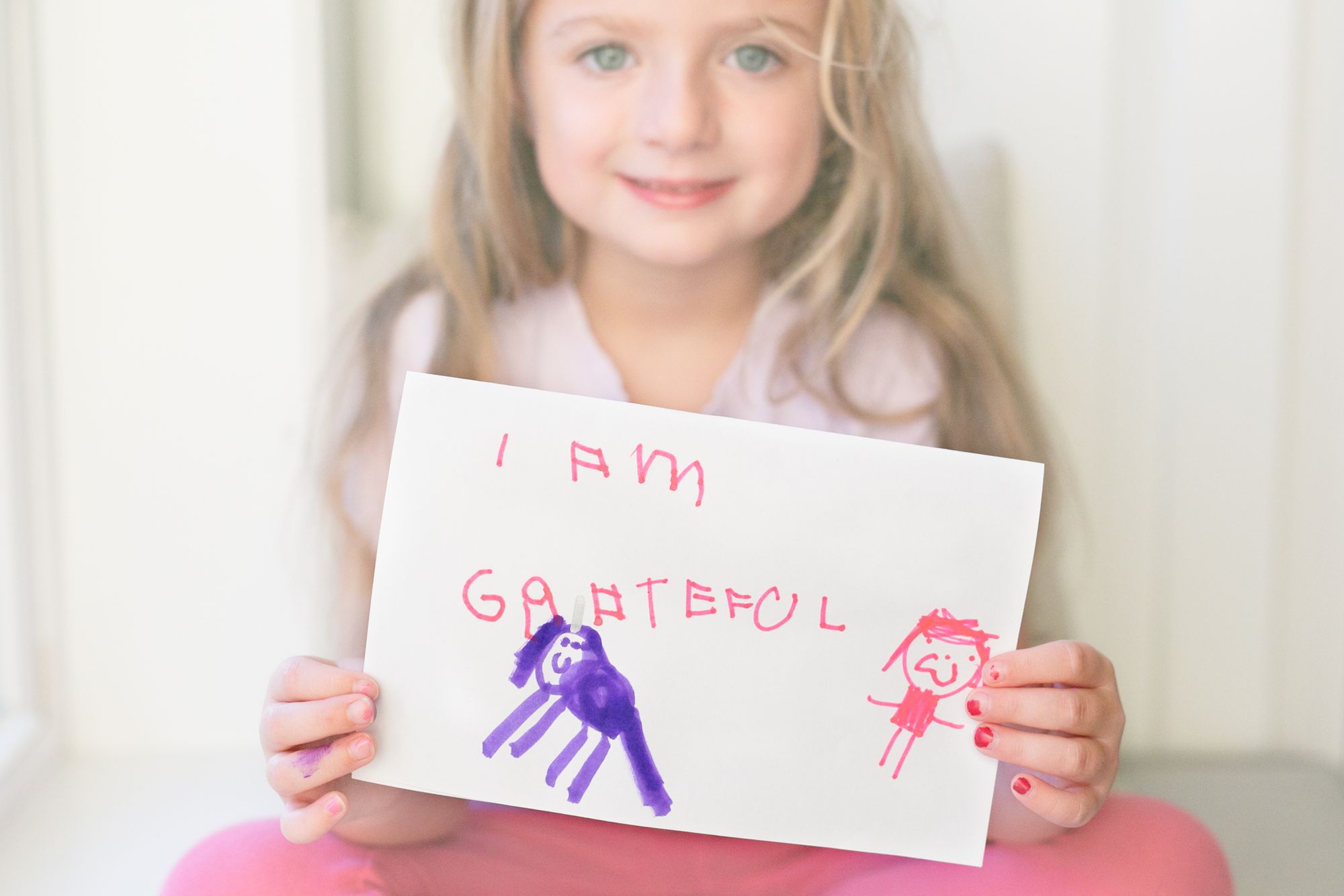 A young girl with blond hair and big blue eyes, the author's daughter, is holding up a hand-made thank you card with the words "I AM GRATEFUL" written in pink marker above a drawing of a purple unicorn and a smiling person.