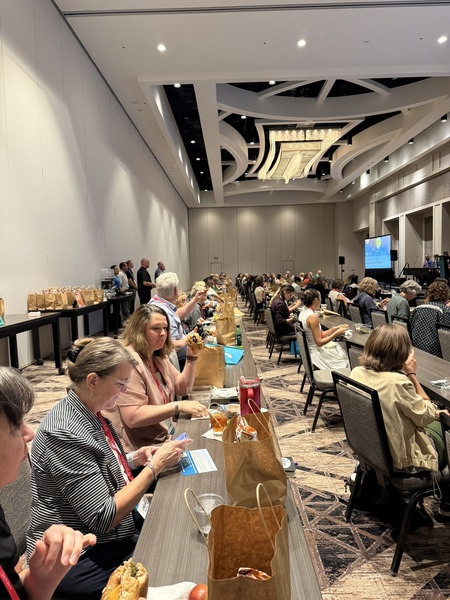 a long wide room crowded with attendees eating lunch during a panel discussion where panelists are raised on a stage next to a projector screen.