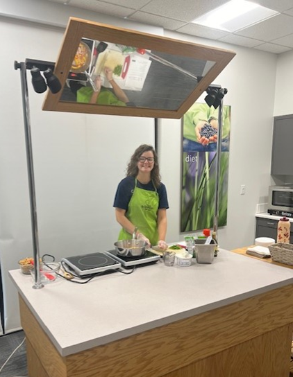 A young woman wearing a blue shirt and neon green apron smiles at the camera. She is standing behind a table that has various ingredients on it as well as two hotplates and a pan. Above the table, angled so that the audience can see, is a mirror that shows what is cooking from a bird's eye view.