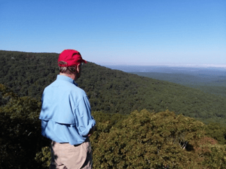 A man wearing a red cap, powder blue sun protection shirt and chinos stands at the top of a mountain vista looking out into the horizon.