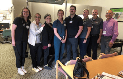 a group of 8 people most dressed in scrubs with medical IDs on stand posing for a picture behind a table.