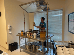 A woman stands in shadow behind a stationary cooking cart that has a mirror angled over-top making what she's cooking on the table easy for an audience to see.