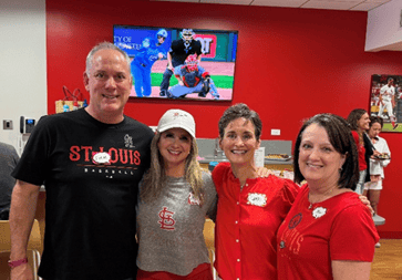 Our Account Management leadership team, one man and three women, pose smiling together in one of the Party Stadiums at Busch Stadium. Behind them is a red wall and a TV showing the game.