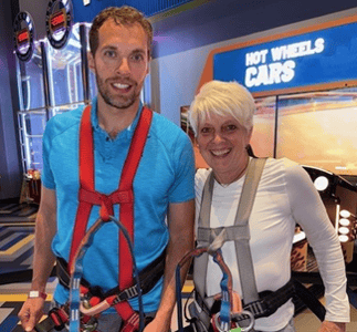A man and woman stand next to eachother wearing harnesses as they prepare to climb an obstacle course. 