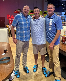 Three Pritikin employees stand together wearing bowling shoes. They are all matching in blue shirts and kaki pants or shorts.