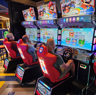 Three men sit in an arcade racing each other playing a Mario game.