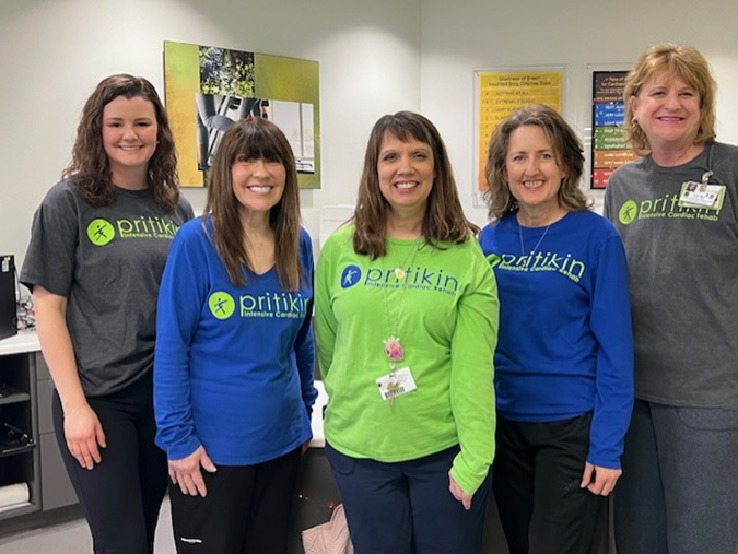 Image of 5 white women wearing Pritikin ICR shirts in grey, blue, and pea green. All are smiling at the camera with their hands at their sides. 