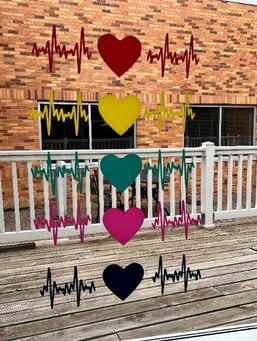 A window with 5 window stickers of hearts and heart rhythm lines next to each. All are different colors. The window overlooks a wooden deck and white fence and beyond that, another building at one of the Centra Care hospitals. 