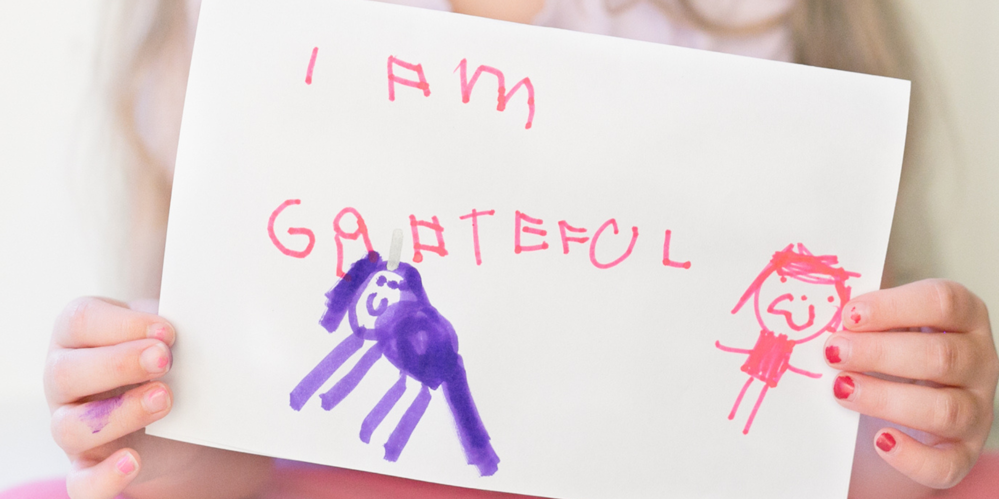 Tiny hands with red chipped nail polish hold a rectangle of white paper. Written in pink marker in a child's handwriting is "I am grateful" . Under the text are two stick people, one in purple marker and the other in pink.