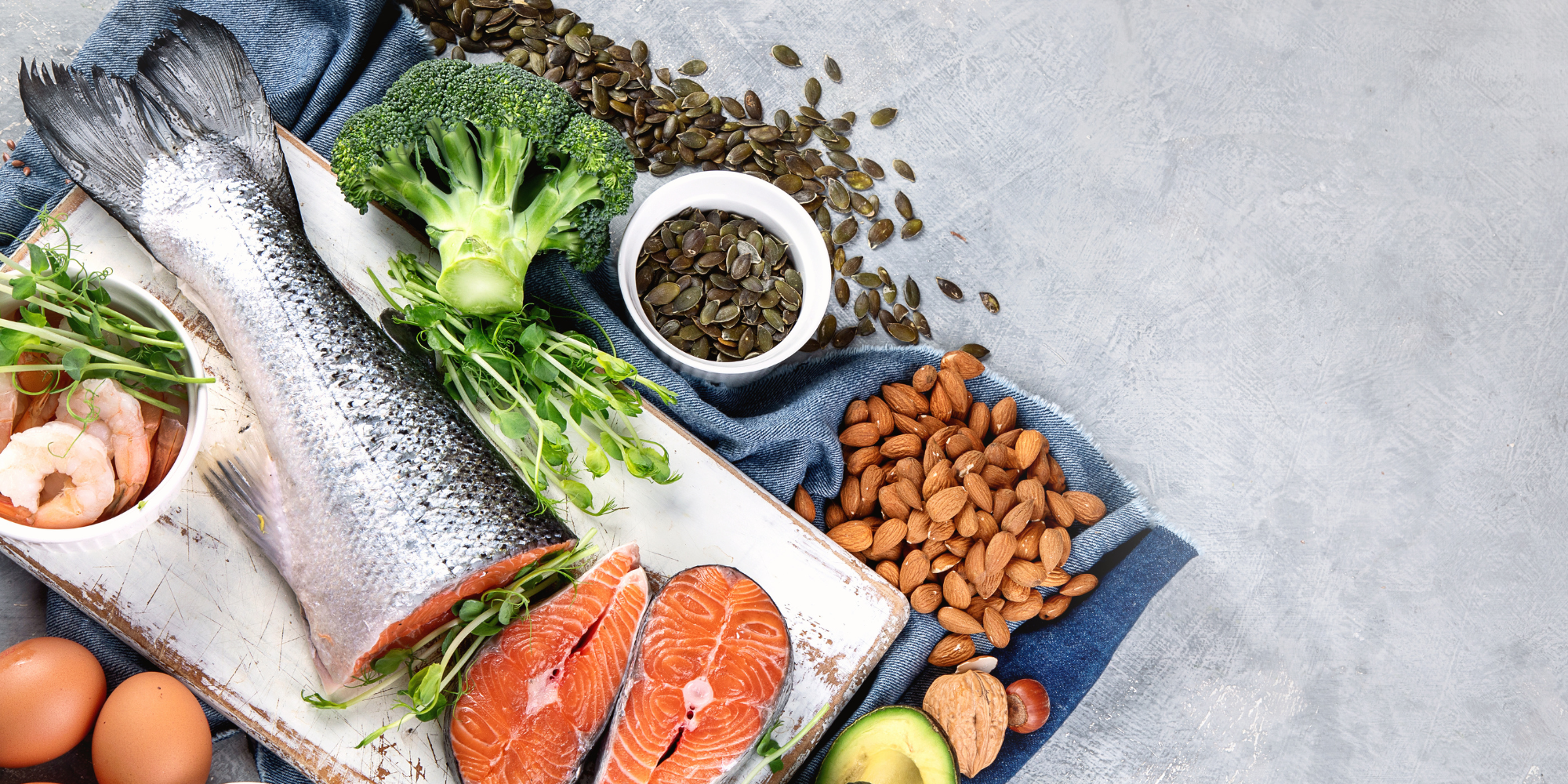 An assortment of omega-3 rich foods on a counter arranged to the left of the image. Laid out is the tail section of salmon along with two salmon steaks, a head of broccoli, almonds, avocado, and pumpkin seeds