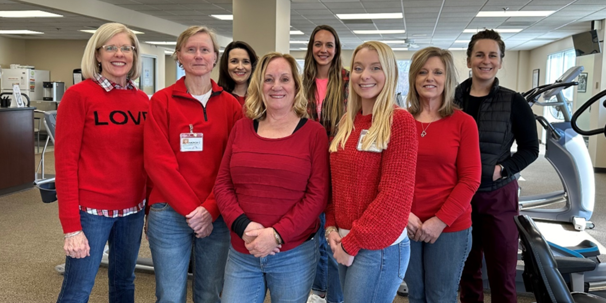 Parkwest team smiles at the camera posing for a group photo in their exercise room. They are all wearing red tops and blue jeans as the photo was taken during heart month on national wear red day.  