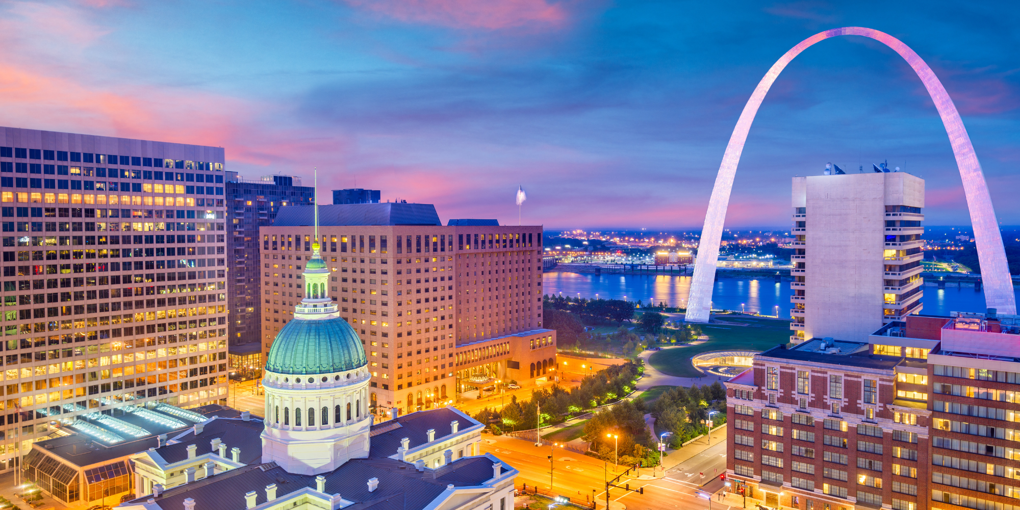 Image of down town St. Louis at twilight. The arch and reflective surface of the Mississippi River beyond are to the right of the image. The city is vibrantly lit up. 