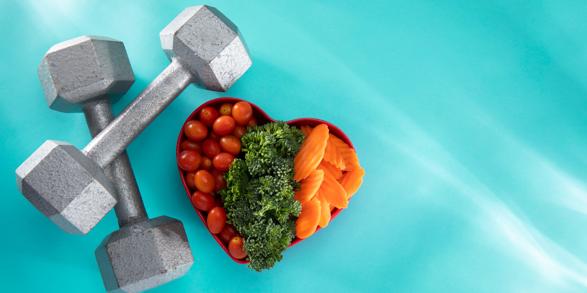 A pair of metal weights (maybe 8 lbs) next to a heart shaped plate full of carrots, broccoli, and cherry tomatoes