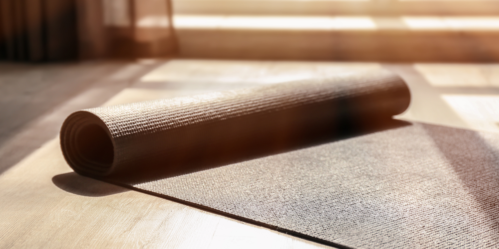 Brown yoga mat unrolled on the floor near a glass door on a sunny day.