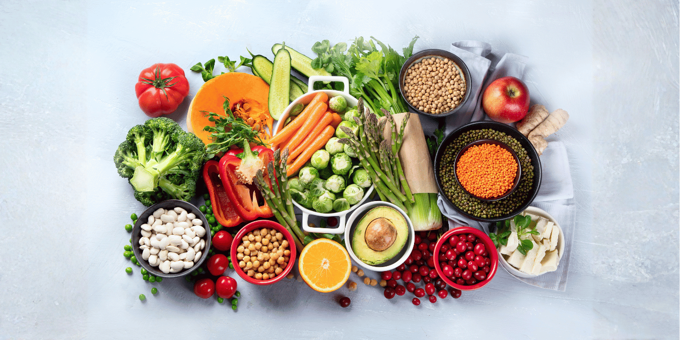 An assortment of fruits, legumes, and vegetables on a white tabletop. The photo was taken from overhead and some items are in bowls while others are loose on the table.