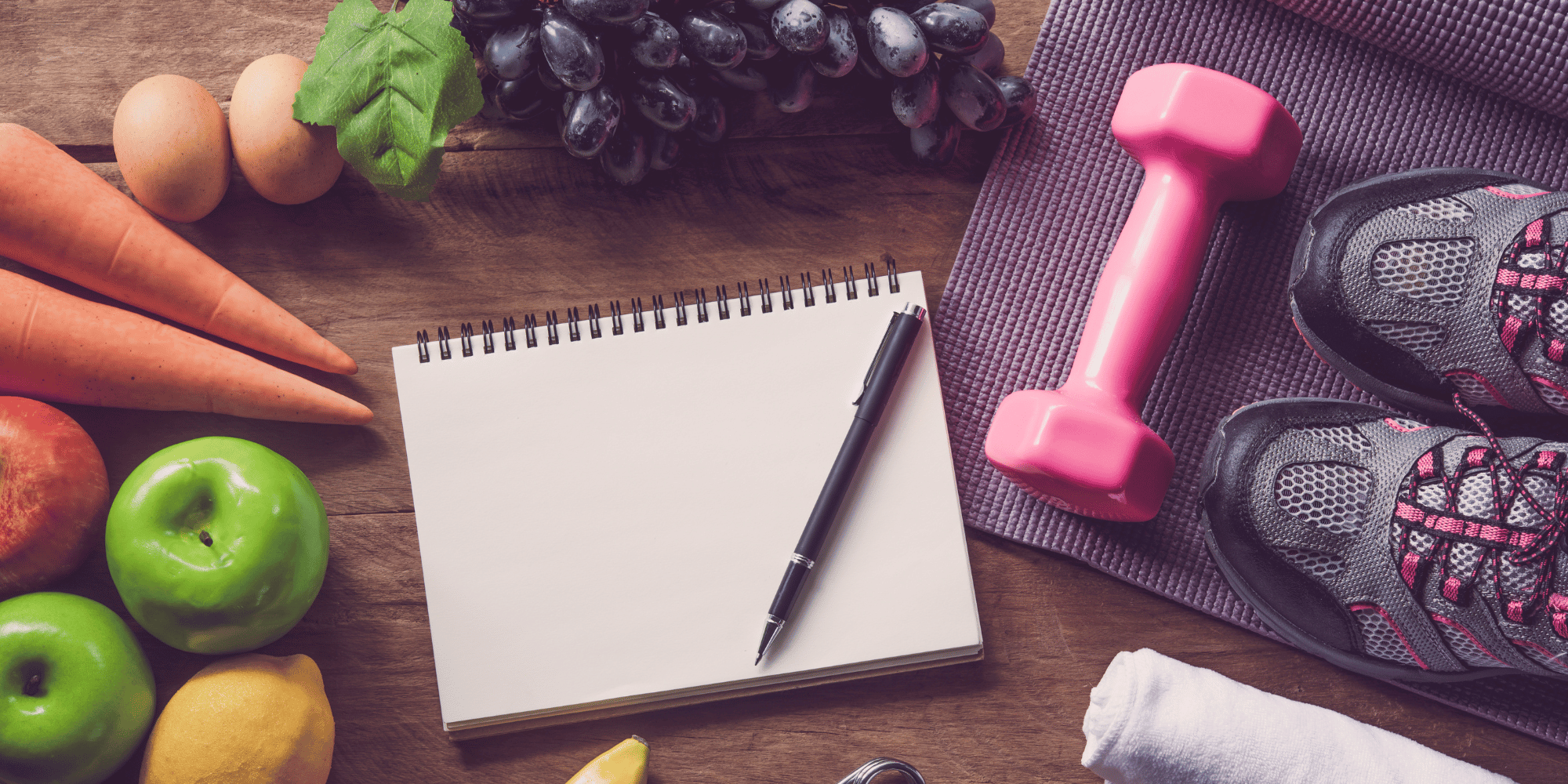 In the center of the image there is a notepad with a pen. All around are fruits and vegetables as well as exercise equipment. 