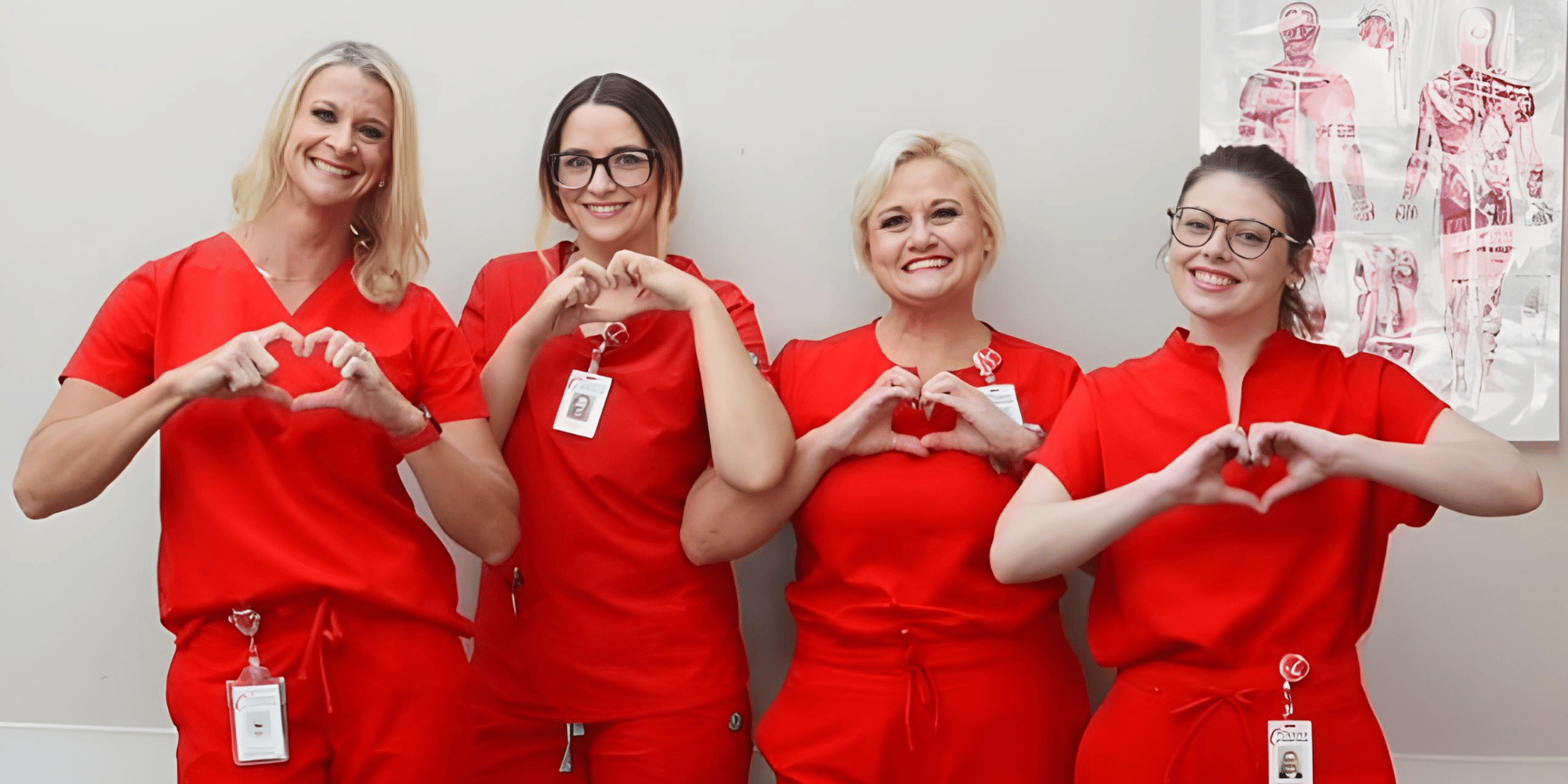 four women wearing red scrubs smile at the camera each making a heart shape with their hands.