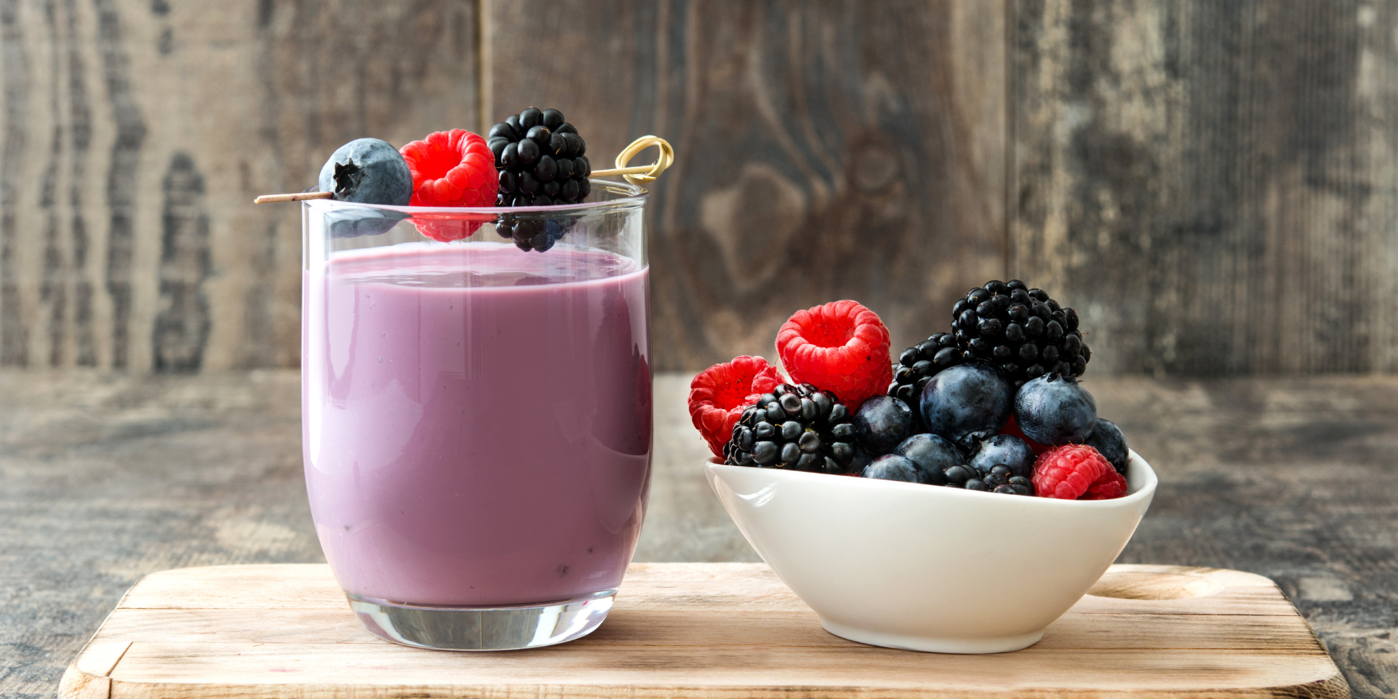 A short glass cup full of a purple berry smoothie. To the right of the glass is a bowl of berries.