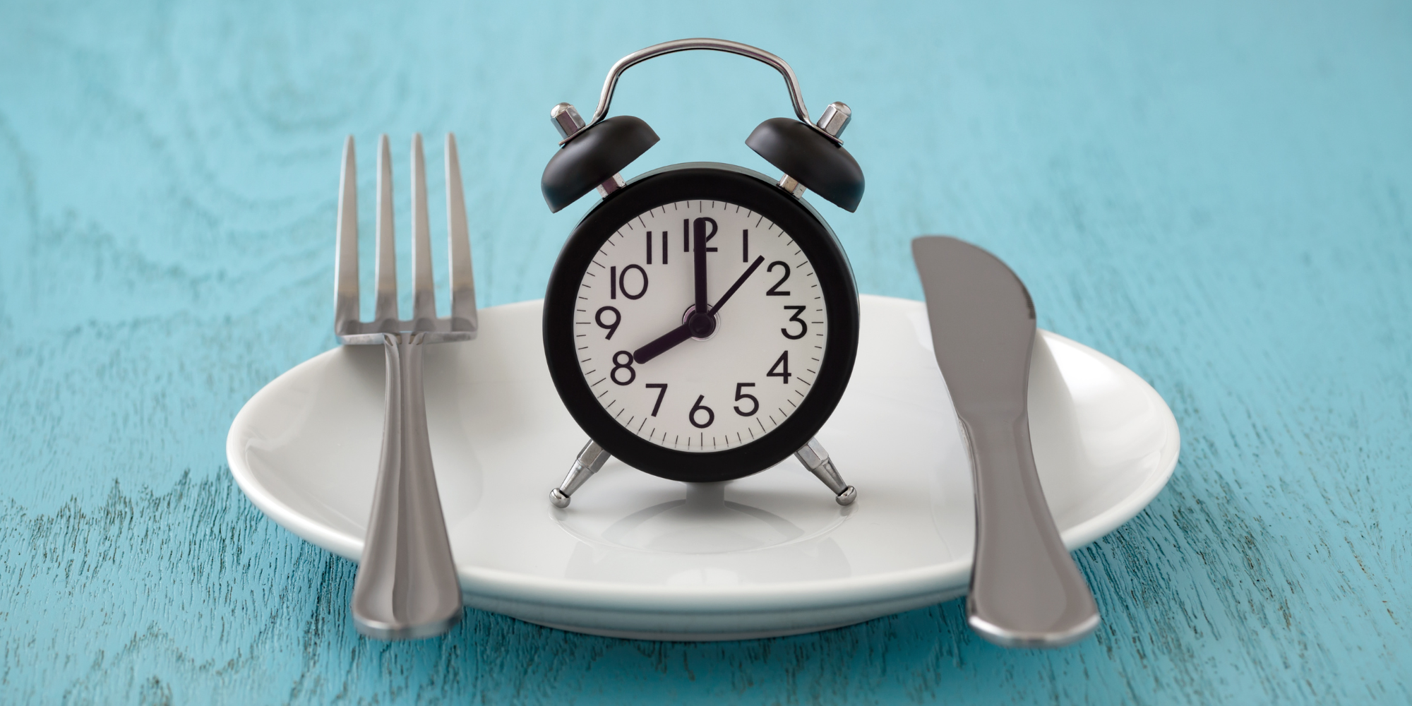 A silver knife and fork are arranged in parallel atop a white plate. Between the utensils, there is an old fashioned analogue alarm clock. The plate has no food on it symbolizing intermittent fasting. 